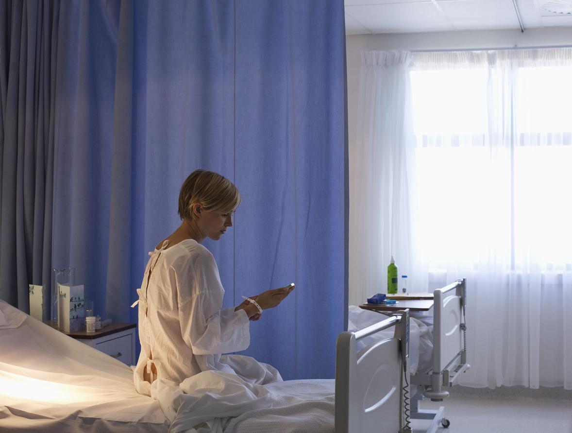 Woman in hospital gown sits on her bed next to a blue privacy curtain to make a phone call