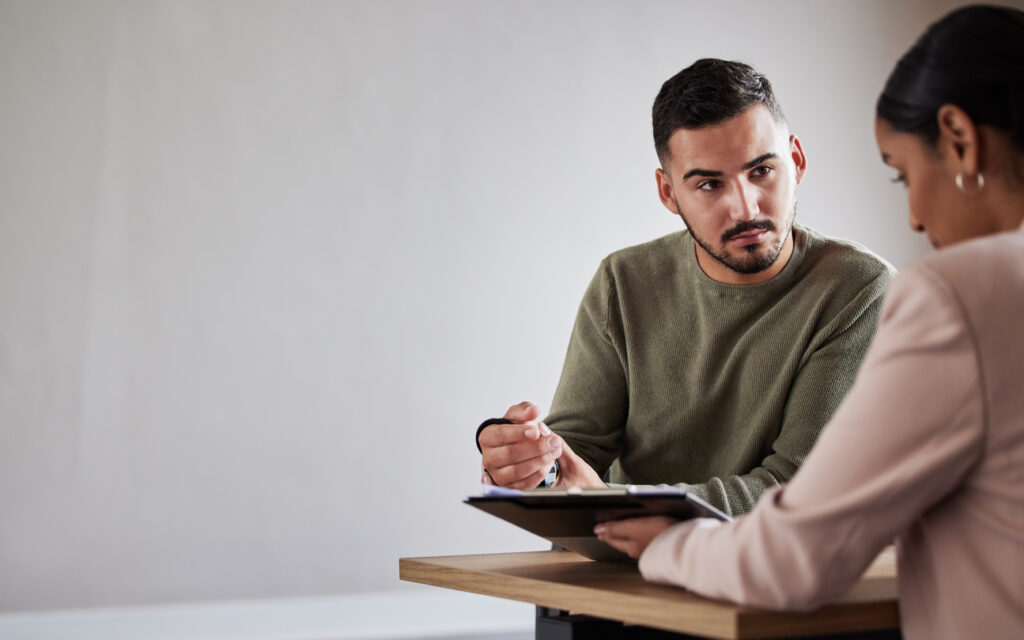 A man sitting at a wooden table with a woman who is showing him forms on a clipboard. The man looks concerned and is holding his injured wrist, which is in a black wrist brace.